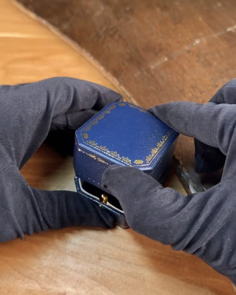Black-gloved hands opening a blue ring box on the atelier bench