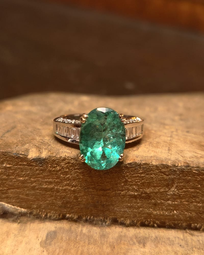 Ring with a large green oval emerald and baguette diamonds along the shank, resting on a wooden bench