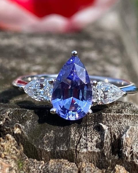 Trilogy ring with a blue-violet pear-shaped stone at the centre and two pear-cut diamonds, photographed outdoors on wood