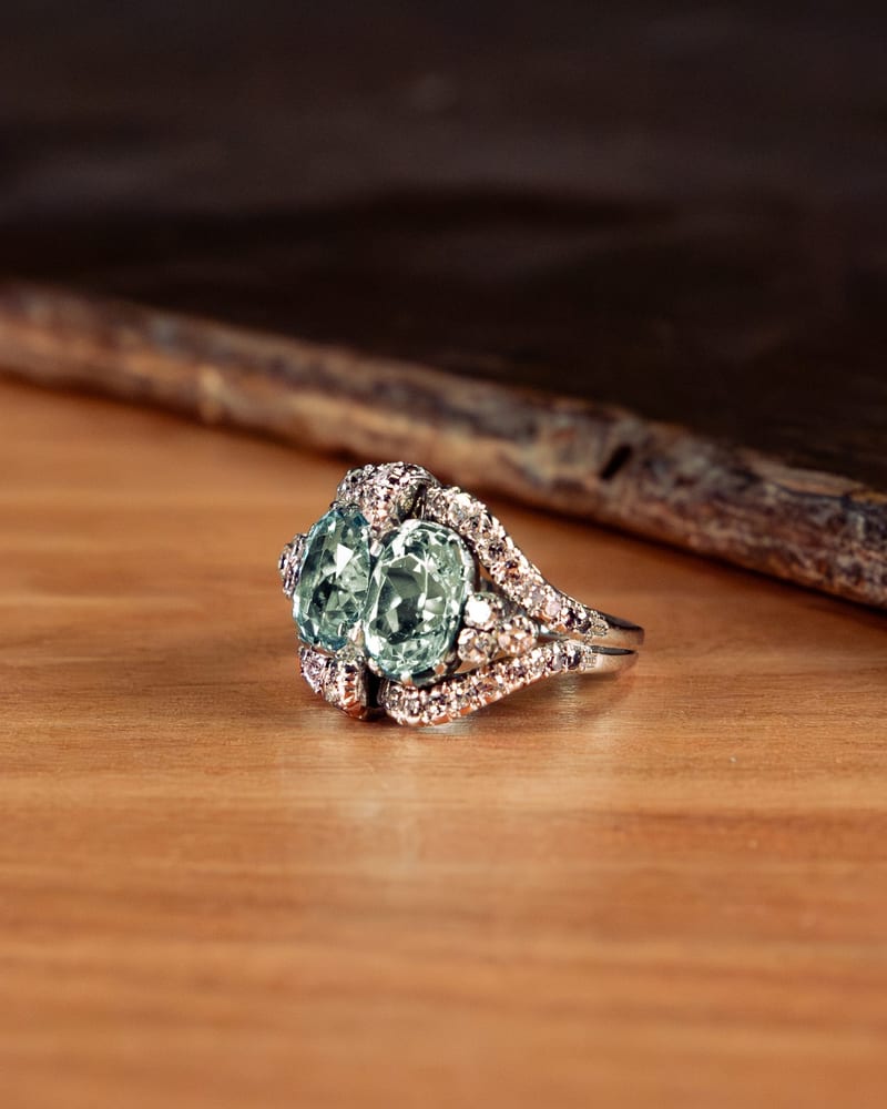 Ring with two blue-green oval stones side by side and pavé diamonds, on a light wooden surface